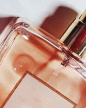 A close-up image of a perfume bottle with a light pink liquid inside. The bottle has a rectangular shape with a gold cap and a white label. The background is soft and light, with subtle shadows creating a gentle, elegant effect.