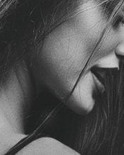 A black-and-white close-up of a person's profile, showing their neck, shoulder, and long hair, with a soft focus on their facial features.