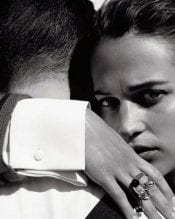 Close-up black and white portrait of a woman looking intensely at the camera while embracing a man in a suit, evoking a sense of intimacy and elegance.