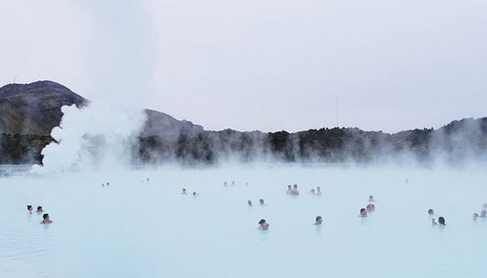 A group of people bathing in a large, misty geothermal hot spring with pale blue water, surrounded by a mountainous landscape and steam rising in the background.
