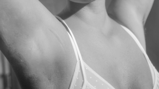 A black-and-white close-up of a person raising their arms, wearing a polka-dot patterned bra, focusing on the upper chest and underarm area.