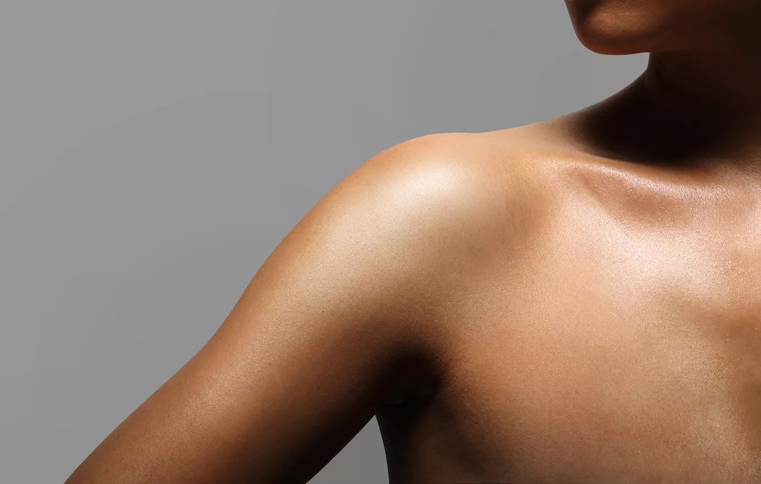 A close-up photograph of a person's shoulder and upper arm against a plain gray background. The skin has a glossy, highlighted finish, emphasizing the contours and smoothness of the shoulder area with soft lighting and shadows.