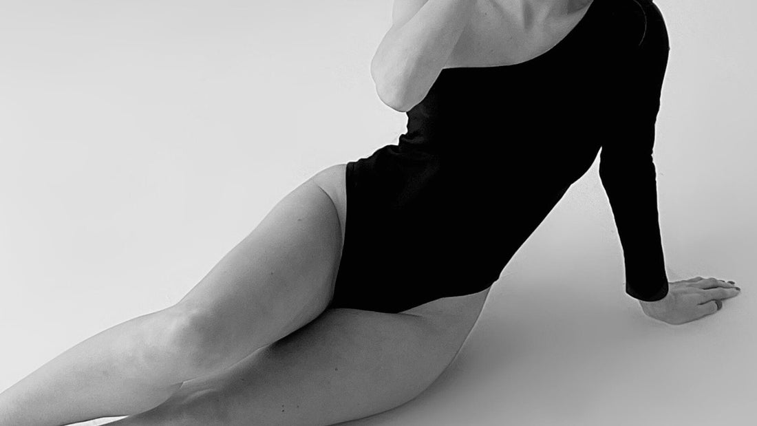 A black-and-white photograph of a person wearing a black long-sleeved bodysuit, reclining on the floor with one leg bent and an arm propped up. The background is plain white, creating a minimalist and high-contrast setting.