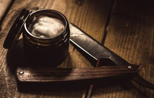 A wooden surface holds a traditional shaving setup: an open dark jar of white shaving cream with its lid beside it, and a closed straight razor with a wooden handle and engraved blade. The scene is softly lit, creating a warm, rustic atmosphere.