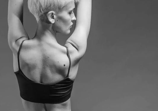 person with a short haircut, viewed from behind, wearing a black sports bra. The person has their arms raised above their head, highlighting the muscles and contours of their back against a plain gray background.