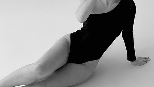 A black-and-white photograph of a person wearing a black long-sleeved bodysuit, reclining on the floor with one leg bent and an arm propped up. The background is plain white, creating a minimalist and high-contrast setting.