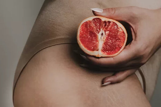 A close-up of a person holding a halved grapefruit in front of their lower body, wearing beige underwear, with visible stretch marks on their skin.