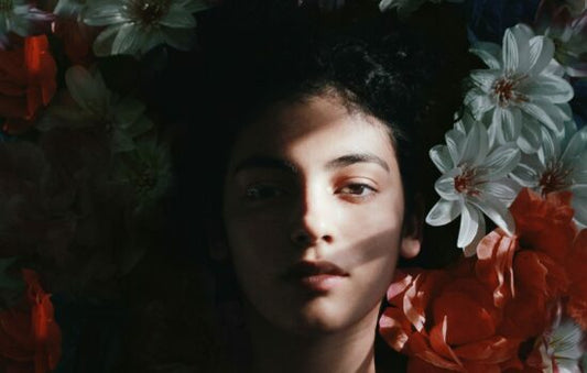 A person with dark hair surrounded by large white and red flowers, with dramatic lighting casting shadows across their face.